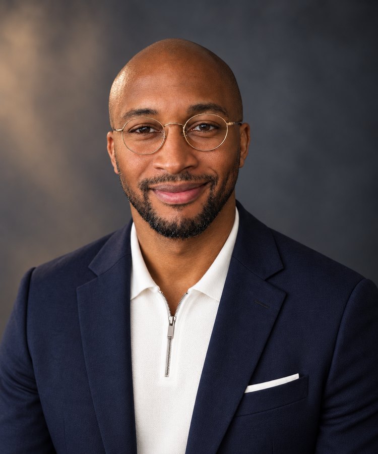 Portrait of Jay Burgess, wearing round glasses, a navy blazer and a white quarter-zip sweater, against a grey backdrop.